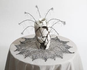 Abstract ceramic sculpture with wire and glass embellishments, displayed on a textured tablecloth.